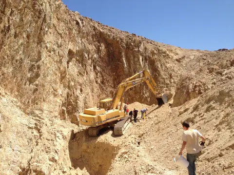 An excavator operates in a rocky ravine while people work around it.