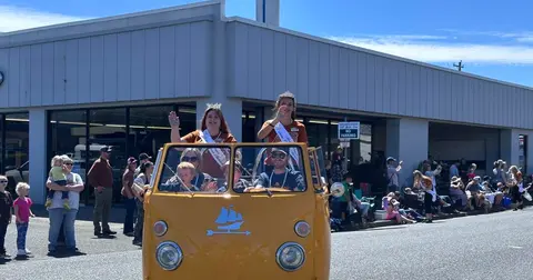 People wave from an orange van during a parade in front of a building.