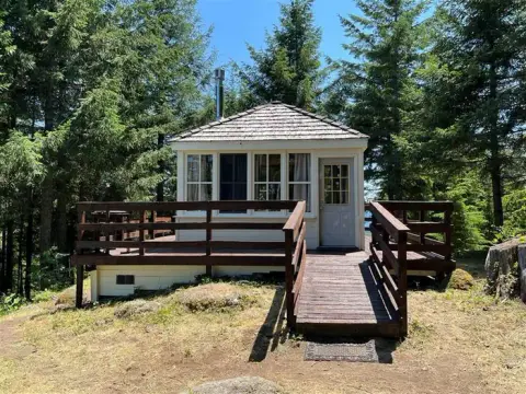 A small cabin with a wooden deck and ramp sits surrounded by tall trees.