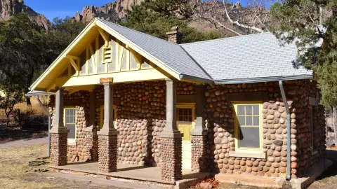 A stone building with a yellow-trimmed porch sits in a landscape with rocky hills.