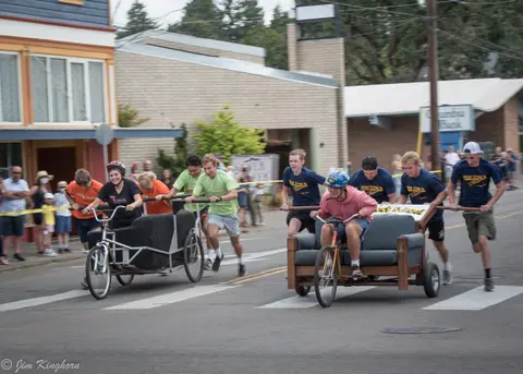 People race custom couch bicycles down a street in a parade event.