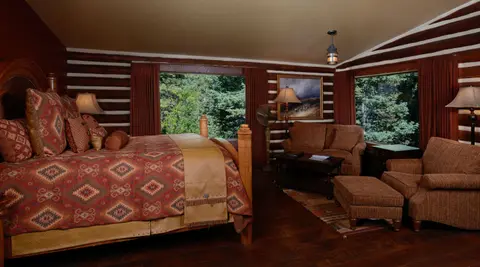 A rustic bedroom with a wooden bed, seating area, and large windows looking outside.