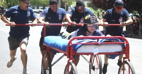 Four people pushing a wheeled bed frame with another person riding it on a street.