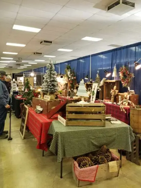 An indoor view of a craft or holiday fair display with tables, decorations, and small Christmas trees.