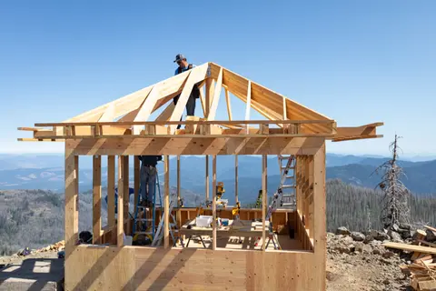 Workers framing the wooden roof of a small building on a hilltop.