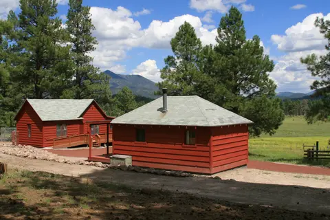 Red wooden cabins stand near a dirt road with trees and mountains in the background.