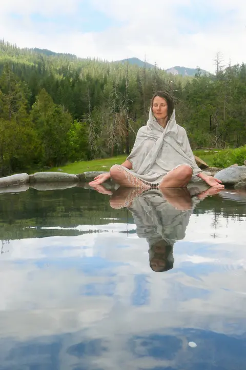 A woman meditates by a tranquil pool with a forest backdrop.