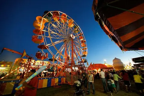 A brightly lit Ferris wheel stands at a crowded fair at dusk.