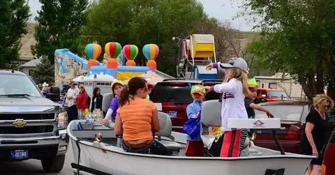 People ride on a float in a parade with carnival attractions in the background.