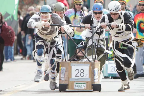 People participate in a costume race, pushing a small wooden cart labeled with the number 28.
