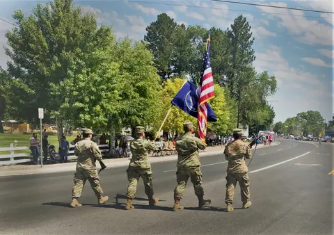 Four soldiers in uniform march down a street carrying flags.