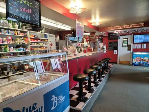 An interior view of a retro diner counter with black stools, checkered flooring, and a stocked snack area.
