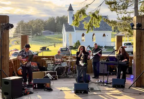 A band performs on an outdoor wooden stage with a white building in the background.