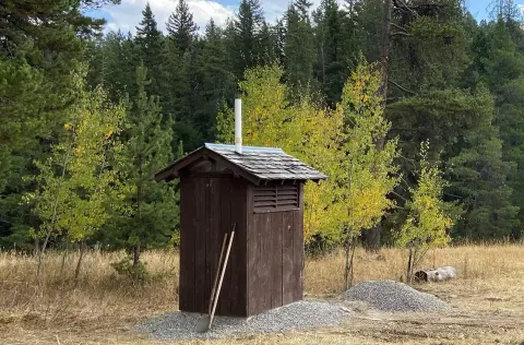 A wooden outhouse stands in a clearing surrounded by trees.