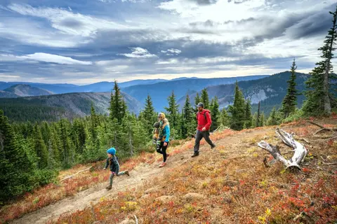 A family hikes along a mountain trail with a scenic view of distant ridges and forest.