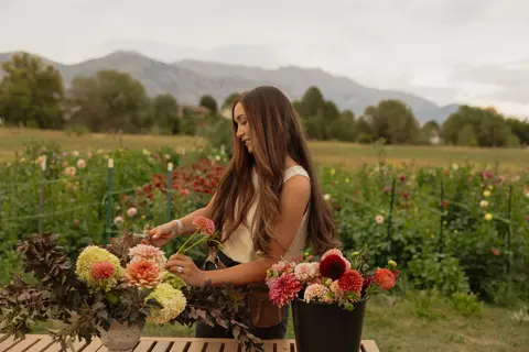A woman arranges flowers outdoors with a mountain range in the background.