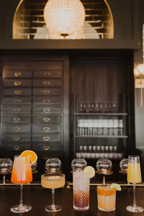 Several colorful cocktails are lined up on a bar counter in front of shelves and a lit fixture.