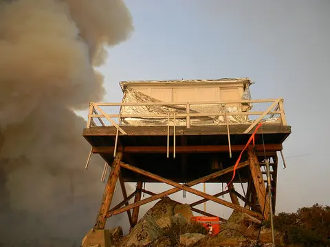 A fire lookout tower is wrapped in protective foil with thick smoke rising in the background.