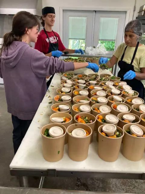 Three people work together in a kitchen preparing containers of food.