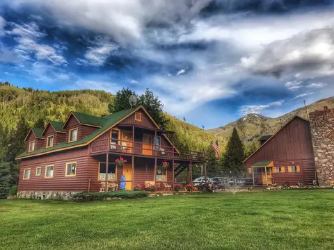A large wooden lodge with a green roof stands in a grassy valley beneath a cloudy sky and mountains.