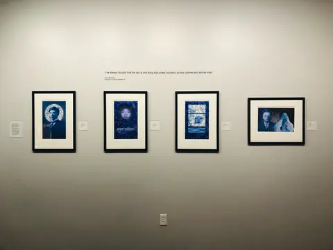 An interior shot shows four framed artworks hanging on a white wall in a gallery.