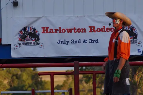 A rodeo clown stands in front of a banner for a rodeo event.