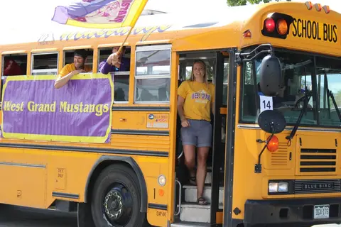 A woman steps down from the open door of a yellow school bus decorated for a parade.