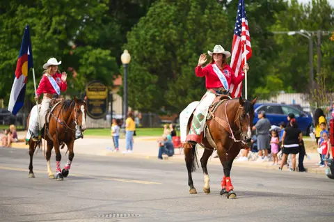 Two women ride horses carrying flags in a parade.