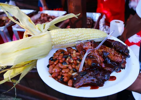 A plate of barbecue ribs, beans, and corn on the cob is held up at an event.