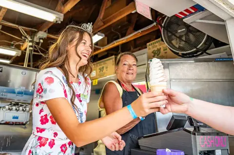 A woman wearing a tiara hands a soft-serve ice cream cone to a customer at an ice cream stand.