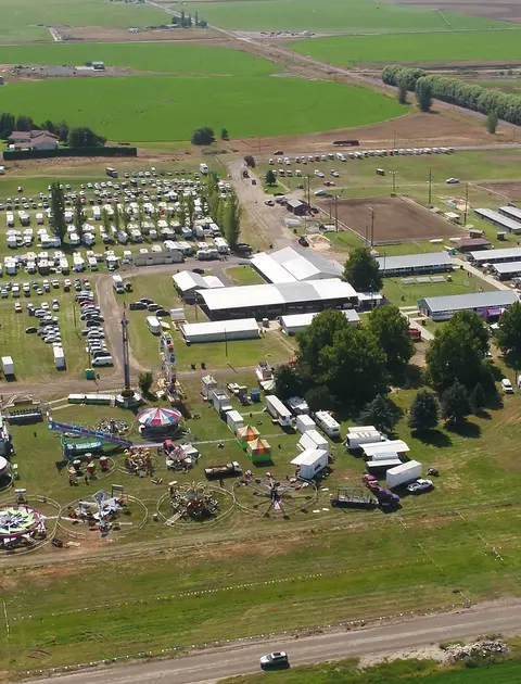 An aerial view shows a fairgrounds with carnival rides, a large parking lot filled with RVs, and surrounding farmland.