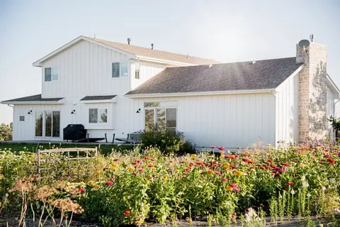 A large white house stands behind a colorful flower garden.