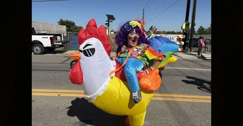A clown rides an inflatable chicken costume in a street.