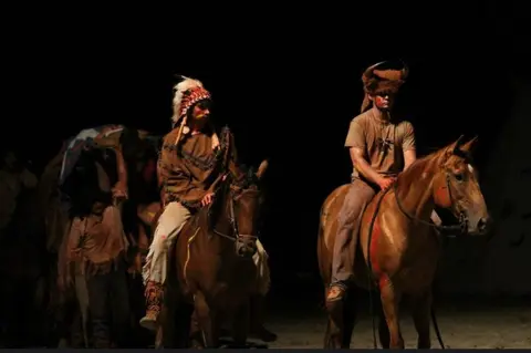 Two people in historical attire ride horses in the dark.