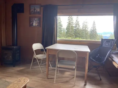 An interior view of a rustic cabin showing a table, chairs, wood stove, and a window overlooking pine trees.