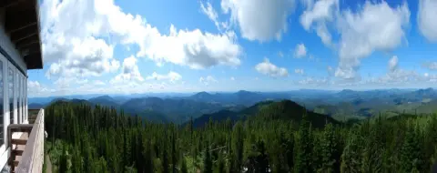 Panoramic view of a forested landscape and distant mountains from a building balcony under a cloudy blue sky.