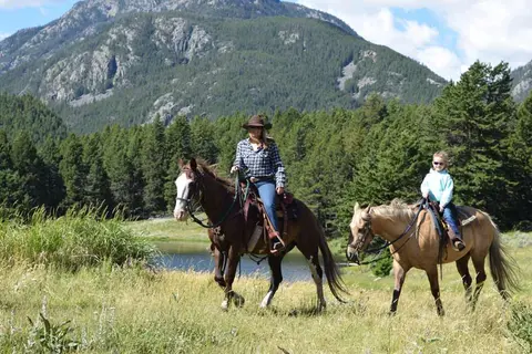 Two people ride horses through a grassy field with a mountain in the background.