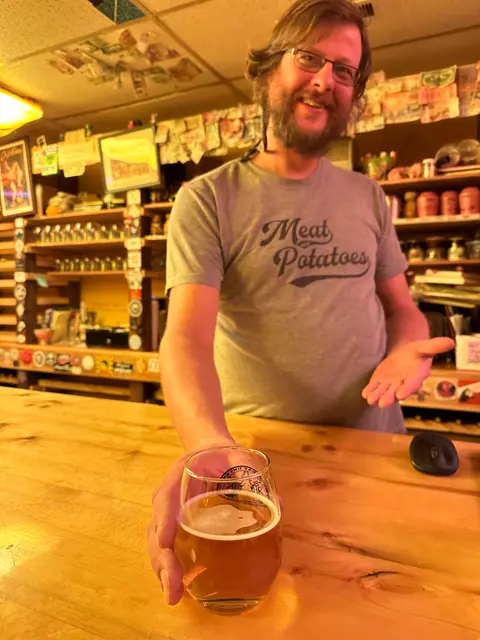 A smiling bartender presents a glass of beer across a wooden bar.