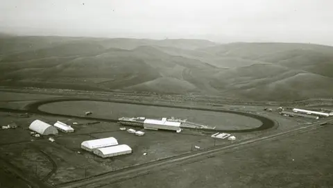 An aerial view shows a racetrack and buildings surrounded by rolling hills.