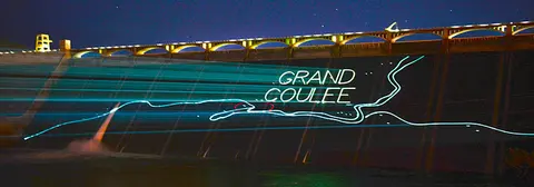 A large dam illuminated at night with laser projections showing text and a map.