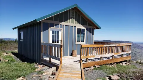 A small cabin with a wooden deck stands on a hillside under a clear blue sky.