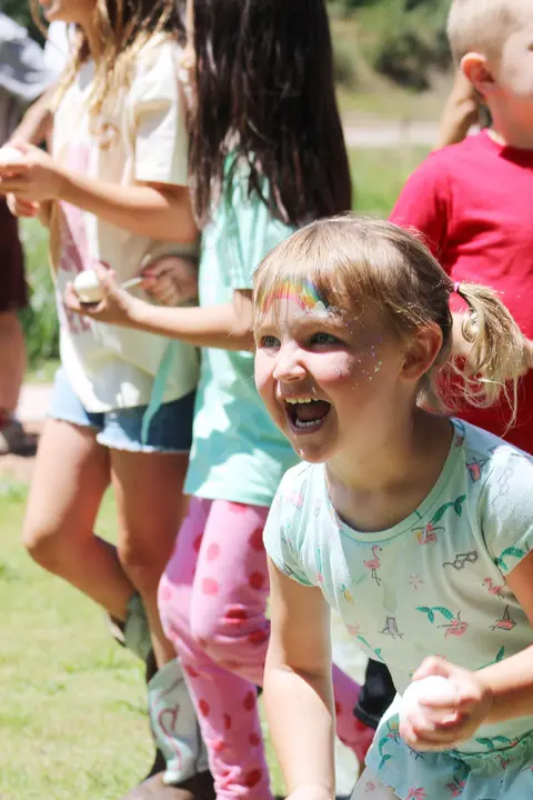 A smiling young girl with face paint holds an egg during an outdoor activity.