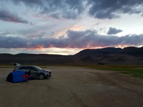 A car parked by a tent in a desert landscape under a cloudy sky at sunset.
