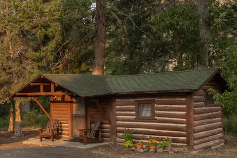 A rustic log cabin stands nestled among trees with two wooden chairs on the porch.