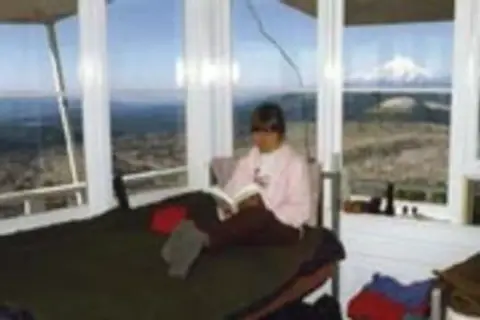 A person reads a book inside a glass-walled tower overlooking a landscape.