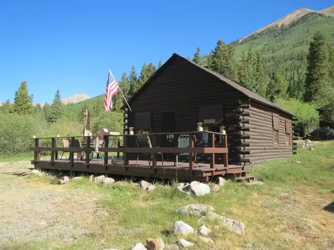 A dark wood cabin with a porch and an American flag stands in a mountain landscape.