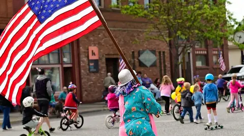 A person dressed in a colorful costume carries an American flag in a parade with children on bikes and skateboards.