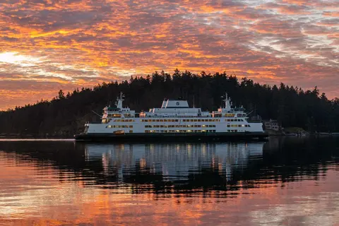 A ferry on calm water at sunset with a tree-lined shore in the background.