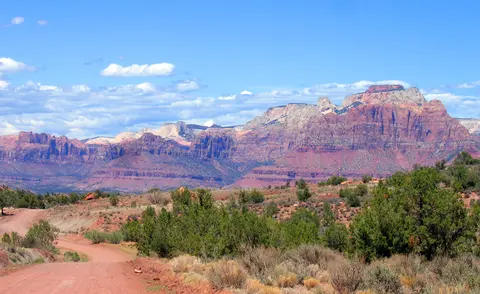 A dirt road winds through a desert landscape towards layered red rock mountains under a blue sky.