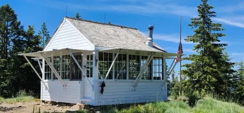 A small white building with many windows and a shingled roof stands surrounded by trees under a blue sky.